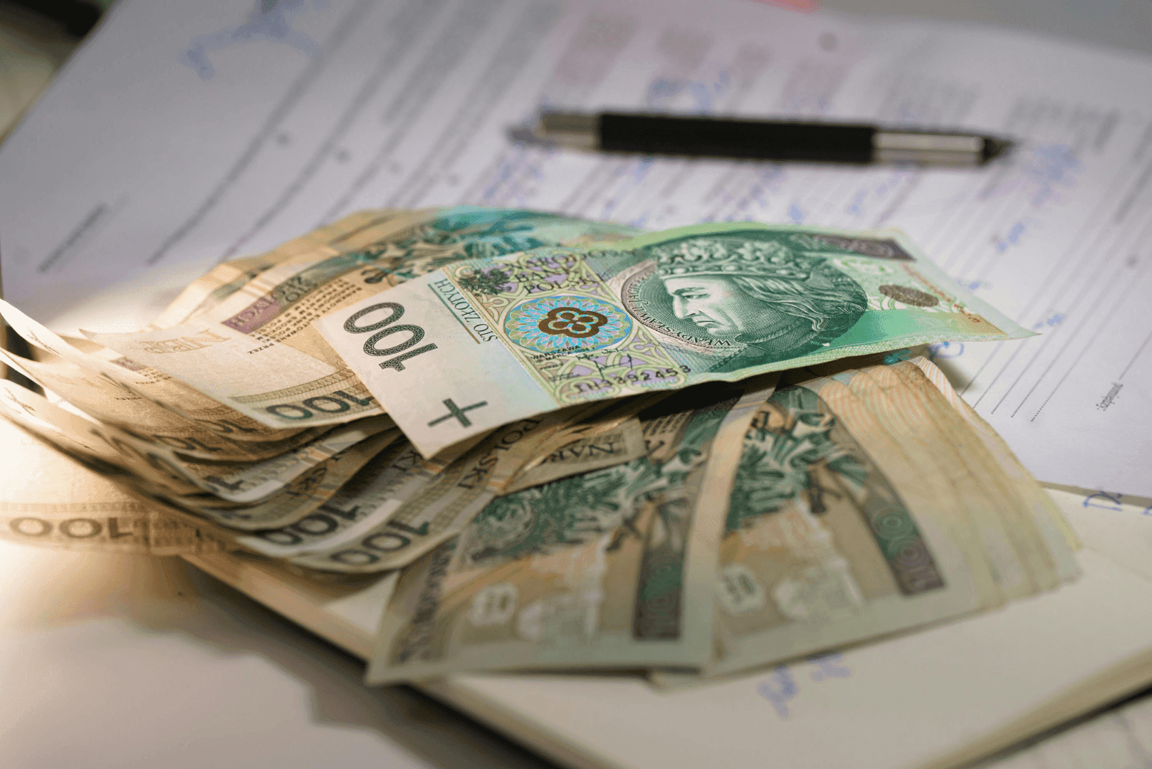 Plik polskich banknotów o różnych nominałach leżący na dokumentach obok czarnego długopisu – ilustracja przewidywalnych kosztów.